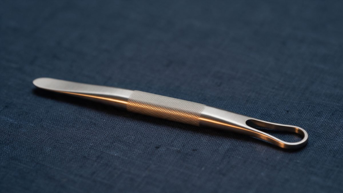 A slender titanium instrument on dark navy linen with a warm amber highlight along its length.