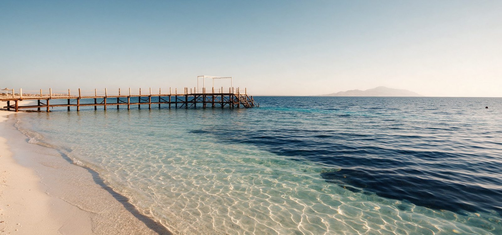 The Red Sea shoreline at Hurghada, glass-clear shallow water over pale sand with a wooden jetty reaching out to sea