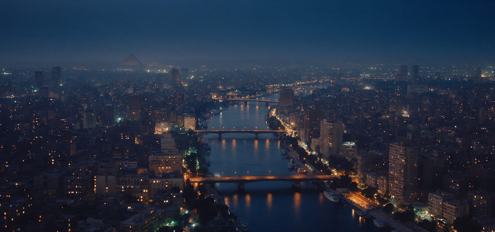 Cairo at blue hour with the Nile threading through the city, lit bridges and warm window lights, the pyramids barely visible in the haze