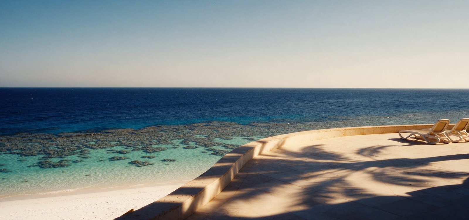 An early-morning terrace above the Red Sea at Sharm El Sheikh, turquoise shallows over the reef and two empty loungers in the palm shade