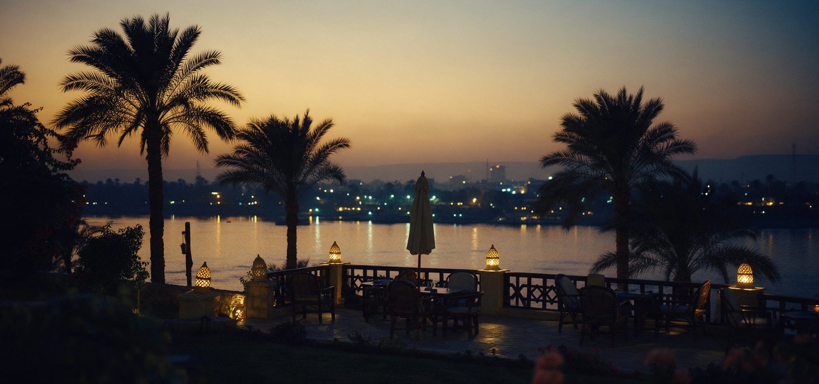A quiet garden terrace above the Nile at dusk, the water reflecting amber light with lanterns glowing and the city skyline soft in the haze